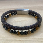 A double-strand beaded bracelet resting on a light wood surface. The top strand is dark brown braided leather, and the bottom strand features smooth  polished tiger eye and black onyx