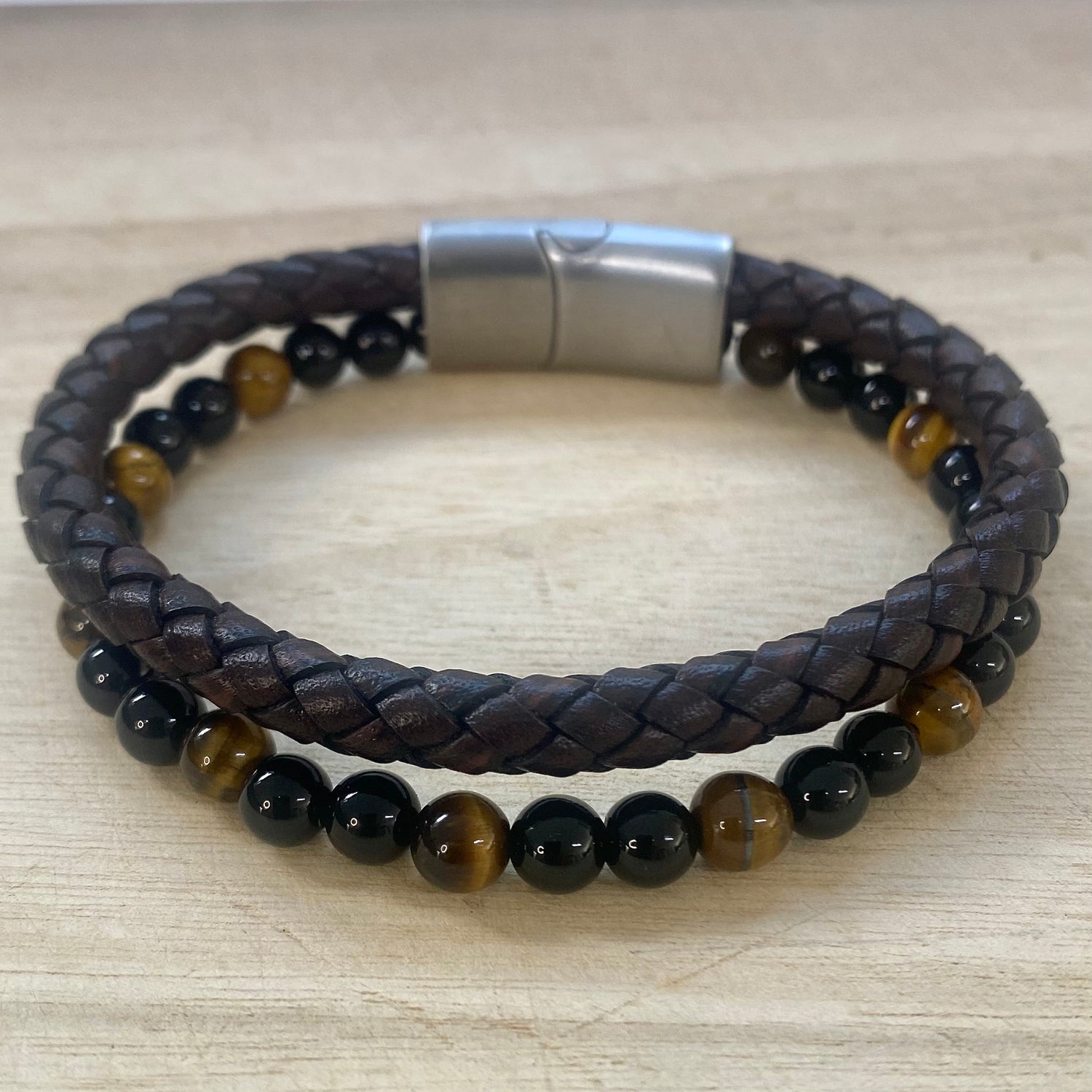A double-strand beaded bracelet resting on a light wood surface. The top strand is dark brown braided leather, and the bottom strand features smooth  polished tiger eye and black onyx