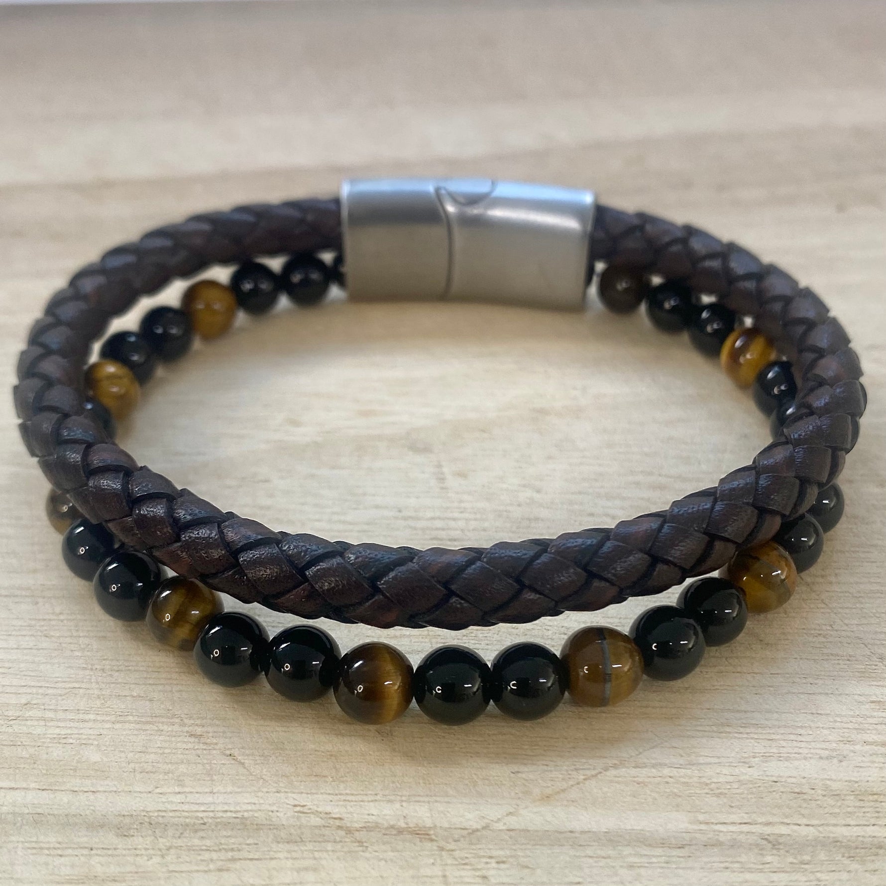 A double-strand beaded bracelet resting on a light wood surface. The top strand is dark brown braided leather, and the bottom strand features smooth  polished tiger eye and black onyx